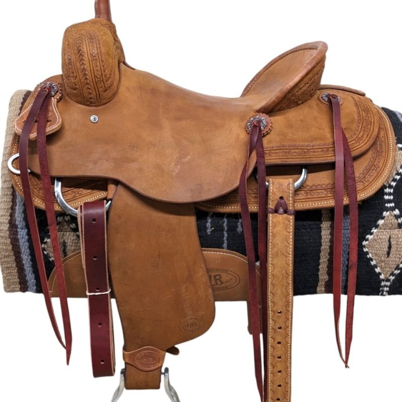 This HR Saddlery ranch cutting saddle is a stunner. This all-roughout saddle is simple which allows the barbed wire border tooling to pop! This saddle has two large, square skirts with rounded edges and saddle strings to keep the rider's gear close at hand. The saddle features a rough-out seat, seat jockey and fenders. Comes with a rear cinch. Weight: Approx. 43 lbs Gullet: 7" mid-concho to mid concho Finished Seat Size: 16" Skirt Length: 28.5" Tree Size: 16.5"